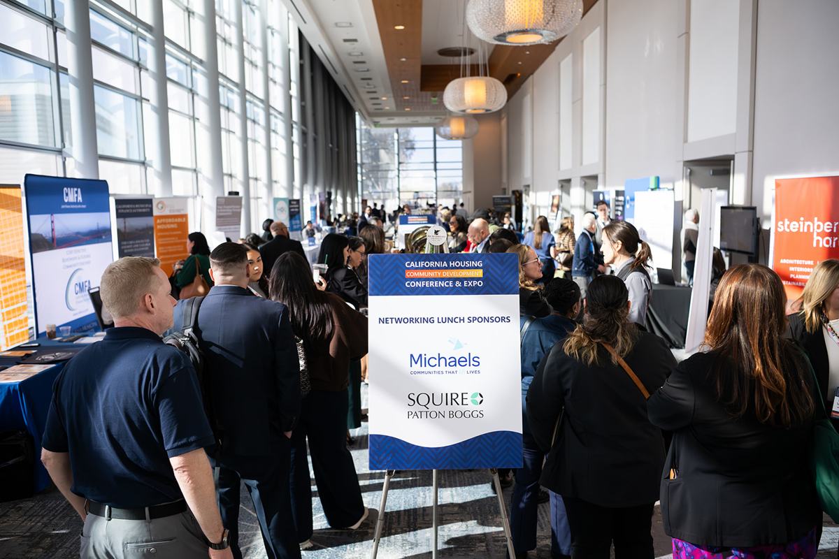 Attendees crowding an exhibit hall beside a sponsor sign